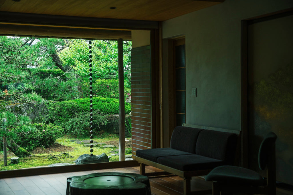 View from a peaceful living room looking at a dark metal rain chain hanging in a lush green garden.